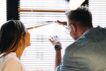 Young cheerful couple looks out the window through the blinds, looks at what is happening outdoor. High quality photoの写真素材