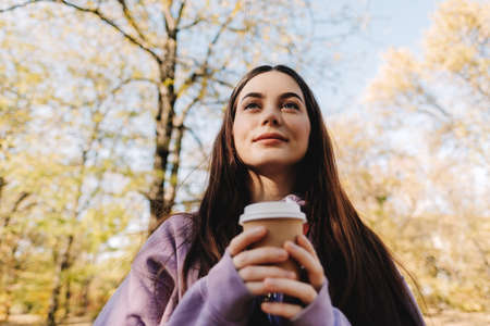 Beautiful young woman drinking coffee and enjoying autumn weather in the park. Woman walking in the...