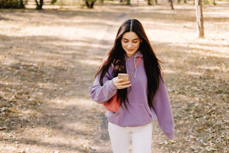 Beautiful young smiling woman using smartphone and enjoying autumn weather in the park. Woman walking in the autumn park. High quality photoの写真素材