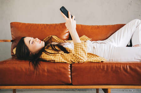 Young calm caucasian stylish woman using smartphone, relaxing lying on comfortable sofa in modern living room, lazy happy woman resting on couch at home. High quality photoの写真素材