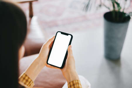 Woman holding a smartphone with a white screen mock up while sitting on the sofa at home. High...
