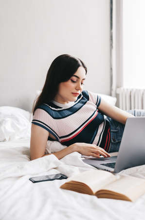 Beautiful young caucasian woman resting on a bed at home and using laptop computer. Chatting with...