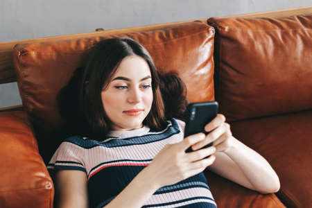 Young calm caucasian woman using mobile phone, relaxing lying on comfortable sofa in modern living room. High quality photoの写真素材
