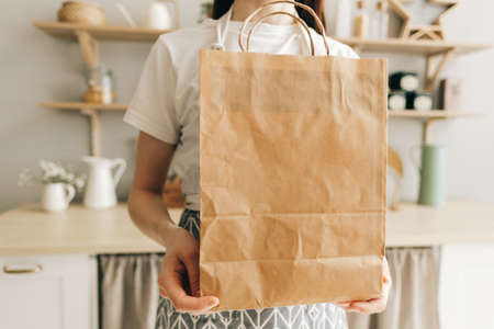 Woman holds eco shopping paper bag with food and vegetables in hands, while standing in modern kitchen. Delivery food, take away.の写真素材