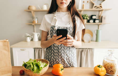 Young caucasian woman in apron use smartphone in the modern kitchen, preparing salad, read recipe. High quality photoの写真素材
