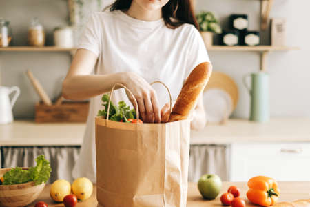 Caucasian woman hold eco shopping bag with fresh vegetables and baguette in modern kitchen. High quality photoの写真素材
