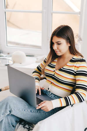 Attractive young caucasian woman use laptop computer, sitting on armchair in bright living room. Distance work conceptの写真素材