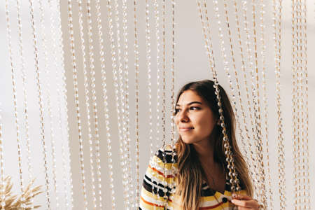 Beautiful smiling young woman in bright living room with white walls and iridescent curtain. High...