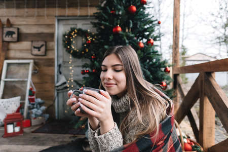 Young caucasian woman in a plaid is drinking a hot drink on the veranda with Christmas tree and decorations.の写真素材