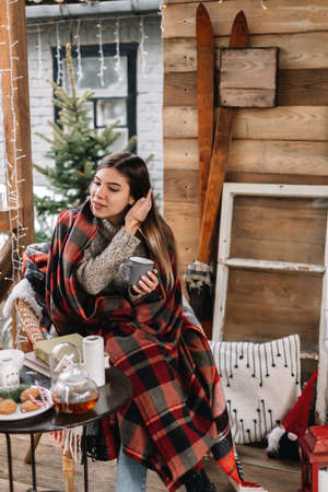 Young caucasian woman in a plaid is drinking a hot drink on the veranda with Christmas tree and decorations.の写真素材