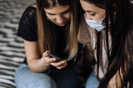 Two young females in medical protective masks at home using a smartphone.の写真素材