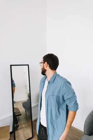 Young caucasian man in glasses standing at the mirror in living room. High quality photoの写真素材
