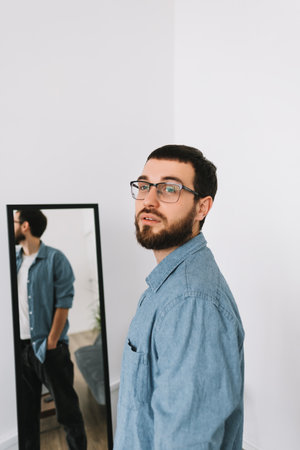 Young caucasian man in glasses standing at the mirror in living room. High quality photoの写真素材