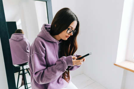 Young stylish caucasian woman using mobile phone near big mirror, sitting on a black chair in white room near window.の写真素材
