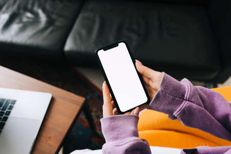 Woman holding a smartphone with a white screen mock up, resting on the armchair in living room at...