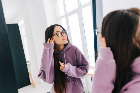 Attractive young caucasian woman in eyeglasses touching face and looking at mirror in living room at home in sunny day.の写真素材