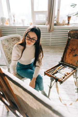 Young caucasian woman artist, holding paintbrush and draws on canvas, working on a home studio.