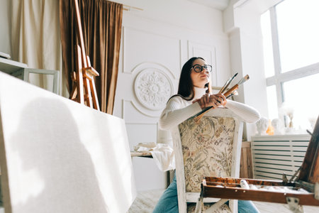 Portrait of beautiful smiling woman artist, sitting on a chair and holding paintbrushes, working on a home studio.の写真素材