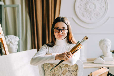 Portrait of beautiful smiling woman artist, sitting on a chair and holding paintbrushes, working on a home studio.の写真素材