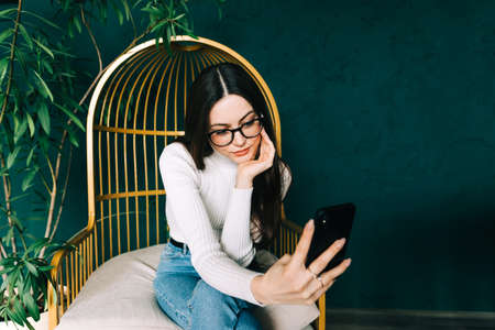 Young caucasian woman holding smartphone looking on cell using mobile phone technology at home, video calling in app virtual chat or shopping online.の写真素材