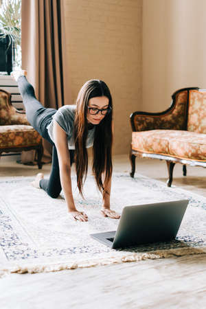 Beautiful brunette fitness woman make stretching exercises in front laptop, doing yoga indoors at...