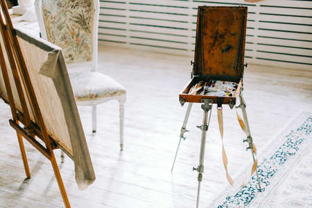 Easel with art paints and brushes in a wooden box at home creative studio.