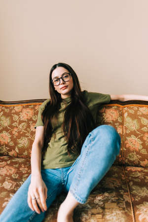Authentic portrait of young caucasian woman in eyeglasses sitting on a sofa at home and looking on camera.の写真素材