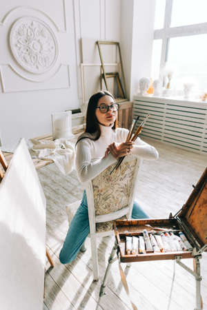 Portrait of beautiful smiling woman artist, sitting on a chair and holding paintbrushes, working on a home studio.の写真素材