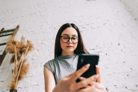 Young caucasian woman in eyeglasses using mobile phone near white brick wall indoor.の写真素材