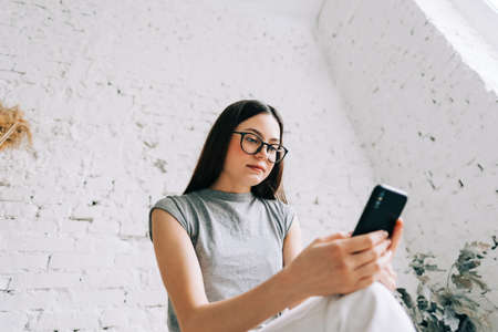 Young caucasian woman in eyeglasses using mobile phone near white brick wall indoor.の写真素材