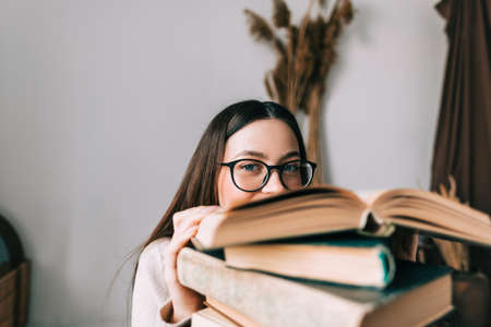 Portrait of young caucasian woman college student in eyeglasses hiding behind a book and looking at...