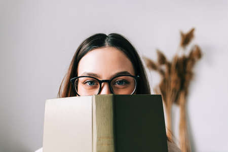 Portrait of young caucasian woman college student in eyeglasses hiding behind a book and looking at camera.の写真素材