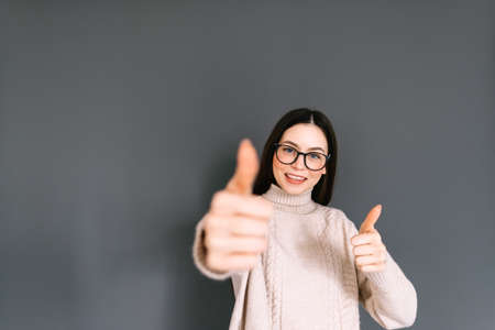 Cheerful caucasian woman in eyeglasses showing thumbs up, doing positive gesture with hand on gray background, looking on camera.の写真素材