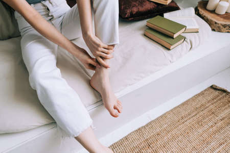 Woman sitting on the bed massages her foot, close-up. Woman with a slender body massages the leg, Young woman massaging her foot on the white bed after training or hard working day. Healthcare conceptの写真素材