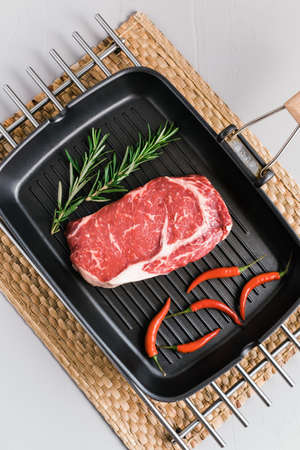 Raw rib eye beef steak on black grilling pan with chili pepper and rosemary, top view.