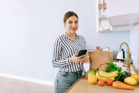 Young smiling caucasian woman use smartphone in the modern kitchen, bag with fresh vegetable on the table. Online buying food and grocery.の写真素材