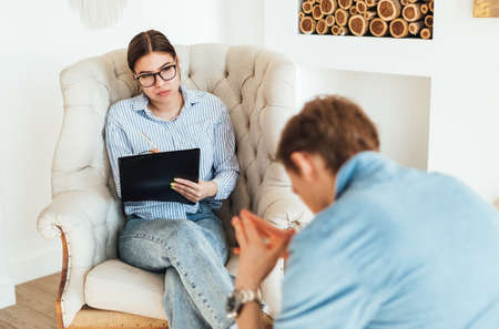 Caucasian woman psychologist in eyeglasses sitting in a chair conducting a psychotherapy session with a patient upset man.の写真素材