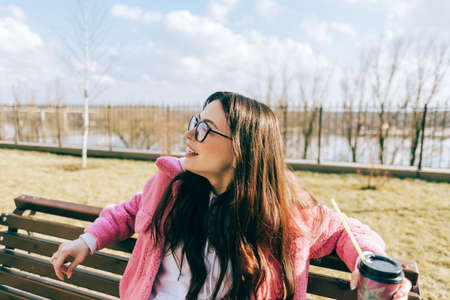 Young caucasian woman sitting on a bench in the park and drinking coffee, enjoying spring weather in sunny day.の写真素材