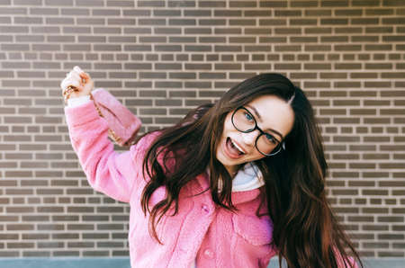 Portrait of young cheerful caucasian woman with brunette hair in eyeglasses and stylish clothing near brick wall outdoor.の写真素材