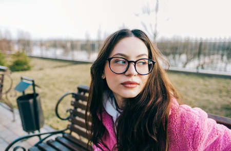 Authentic portrait of young caucasian woman with brunette hair in eyeglasses sitting on a bench in the park.の写真素材
