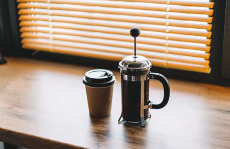 Coffee brewed in a french press and paper cup on a wooden table near window.の写真素材