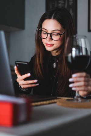 Young caucasian woman with brunette hair resting at home, drinking wine and using smartphone and laptop. Millennial lifestyleの写真素材