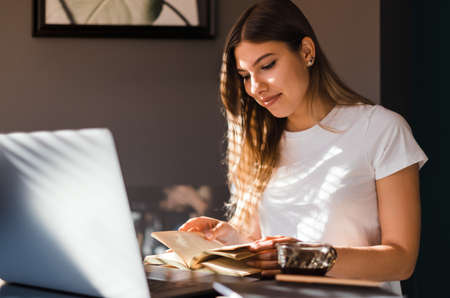 Happy young caucasian woman reading book in front laptop at the kitchen in sunny morning and drinking coffee.の写真素材