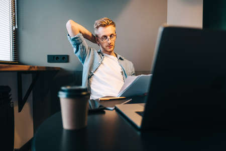 Pensive caucasian man working with laptop at home office and looking on paper documents, calculating bills.の写真素材