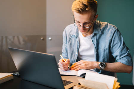 Caucasian man college student in glasses studying with laptop distantly preparing for test exam writing essay doing homework at home, distantly education concept.の写真素材