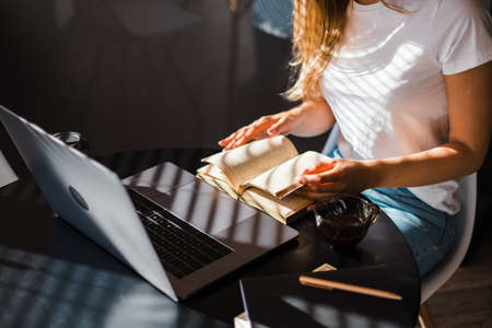 Caucasian woman reading book in front laptop at the kitchen in sunny morning and drinking coffee.の写真素材