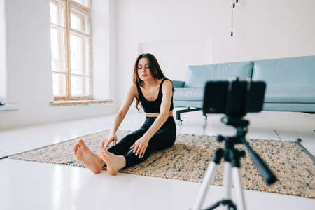 Beautiful brunette fitness woman make stretching exercises in front camera on tripod, doing yoga...