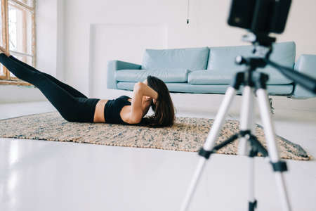 Beautiful brunette fitness woman make stretching exercises in front camera on tripod, doing yoga indoors at home. Staying fit and healthyの写真素材