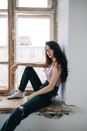 Attractive stylish caucasian woman with long brunette hair sitting on windowsill in living room.
