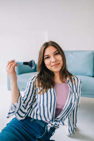 Portrait of attractive caucasian young woman holding makeup brush in hand sitting on floor near blue sofa.の写真素材